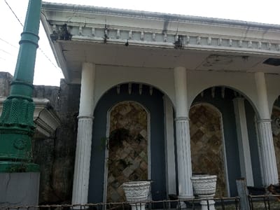 A weathered building exterior features a row of arches supported by fluted columns. The archways reveal stone patterned walls. A decorative cornice lines the roofline, showing signs of age and discoloration. A tall green lamppost stands to the left of the structure. White ornamental planters sit in front, adding a touch of faded grandeur. A partial view of a fence is also seen at the front of the building.