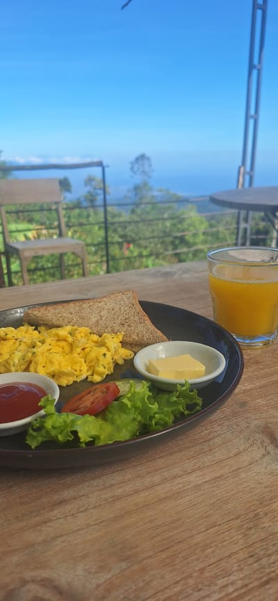 A breakfast plate with scrambled eggs, toast, a small dish of ketchup, a slice of tomato, lettuce, and a pat of butter sits on a wooden table. To the right, a glass of orange juice is visible. In the background, a chair and a table are set against a railing overlooking a lush, green landscape with a distant view of the ocean under a bright blue sky. The scene appears to be in a restaurant or cafe with an outdoor seating area. The exact location within Banjar, Indonesia, is not specified in the image.