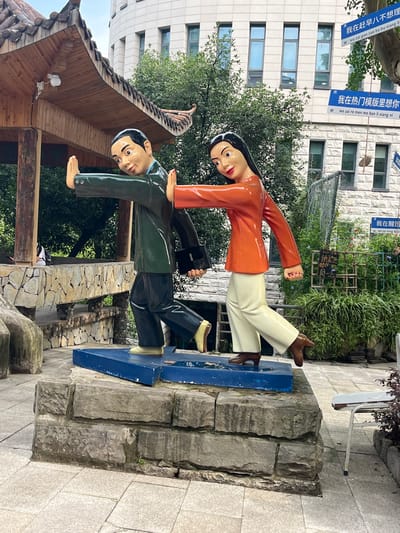 In the Jiulongpo District of China, a public art installation depicts a stylized human figure wearing a dark green suit and black pants, with a briefcase in hand. Another figure, dressed in an orange top and cream-colored pants, is positioned facing the first figure. Both figures are shown with their hands extended, palms outward, as if in a gesture of stopping or pushing away. They stand on a blue, arrow-shaped base, which is mounted atop a layered stone pedestal. The pedestal is situated on a paved area with stone tiling. In the background, a traditional-style wooden structure with a curved roof is visible, along with lush green trees. Further in the distance, a modern building with multiple windows and a white facade is present, and blue signs with Chinese text are affixed to a tree. The overall scene suggests a narrative of human interaction or social commentary, presented in a public outdoor space.