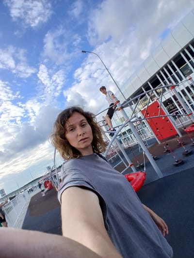 A woman in a grey t-shirt is taking a selfie at an outdoor playground with a child on a climbing structure in the background. The playground has red and grey climbing equipment, and the ground is a dark blue rubber surface. Behind the playground, there is a modern white building with glass windows, a street with cars, and a bridge. The sky is blue with white clouds. The location is the Vorsha, Russia.