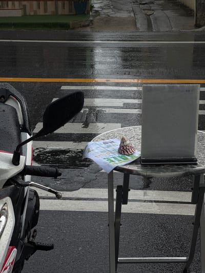 The image shows a street scene in Karon, Thailand, during rainfall. The foreground features a parked motorcycle on the left, with its mirror visible. To the right, a metal table with a glass top sits on a crosswalk. On the table, there is a folded pamphlet and a conical shell decoration. A grey rectangular object, possibly a menu holder or sign, stands upright on the table. The road surface is wet, reflecting the surroundings, and visible raindrops are present in the air and on surfaces. In the background, the wet asphalt road continues, with a sidewalk and building elements visible further back. The scene conveys a quiet, wet day in a Thai city.