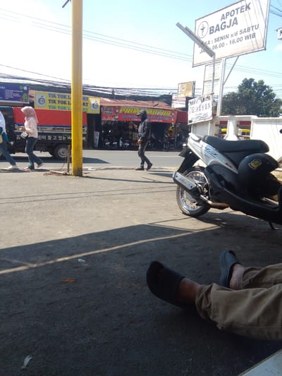In the city of Cianjur, Indonesia, on a sunny day, a street scene unfolds. A bright yellow pole stands prominently in the foreground, casting a shadow on the asphalt road. To the left, a red truck with black siding is parked, displaying signage with Korean text and a logo of a woman with a headscarf. Next to the truck, a woman in a pink headscarf and white shirt walks, holding a white purse and a mobile phone. A man in a black jacket and pants is also walking, his back to the camera. A silver motorcycle with a black seat and helmet rests on the side of the road. In the background, shops and businesses line the street, with a prominent sign for "APOTEK BAGJA" and another for "MOTOR PARK & MIKA MOTOR". The sky is clear and blue. In the lower right corner, a person's legs are visible, wearing brown pants and black slides, resting on the ground.