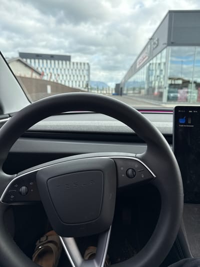 The image shows the interior of a Tesla vehicle parked outside a building complex in Harstad, Norway. The steering wheel, dashboard, and a portion of the touchscreen display are visible. The touchscreen displays "PRND" and "79%" battery life, along with a message in Norwegian: "Trykk inn bremsem." This translates to "Press the brake." Outside the vehicle, a modern building with a distinctive checkered facade is seen in the background, alongside a long, glass-fronted commercial building. The sky is overcast with clouds. A pair of sandals is visible on the floor of the car.