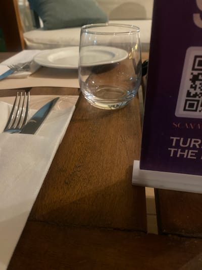 This image shows a close-up view of a table setting with a glass of water, silverware, and a menu. The subjects are primarily objects arranged on a wooden table. The scene appears to be indoors, possibly in a restaurant or dining area.
The activity taking place is a meal service or preparation for one. A clear glass is filled with water, and a fork and knife are neatly placed on a white napkin, suggesting readiness for dining. The menu, partially visible, adds to the context of a place where food is served.
Notable details include the texture of the wooden table, which appears to be rustic, and the clean, modern presentation of the tableware. The lighting is soft, hinting at an indoor environment, and the time of day is not explicitly discernible. The visible text on the menu is "SCANS" and "THE R," suggesting it's a promotional or informational item. The overall impression is one of organized hospitality.
The location context provided is Fagge A, Nigeria. While this information is external to the visual content of the image, it helps to contextualize the setting as likely a dining establishment within that region.