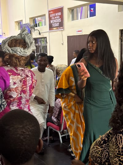 A gathering of people is observed in Lugbe, Nigeria. The setting appears to be indoors, likely a religious space, given the banner displaying the text "GOD'S WORD STILL WORKS WONDERS TODAY."

Individuals of varying ages are present, engaging in social interaction. A woman in a green dress is operating a pink mobile phone. Others are wearing traditional African attire. A man in a white outfit appears to be either sitting or lying down. The lighting is moderate, suggesting an interior environment. The scene seems to capture a moment within an ongoing event.