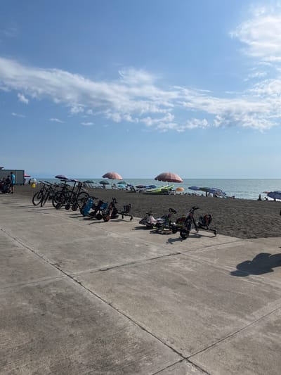 The image depicts a bright, sunny day at a beach in Ureki, Georgia. The scene is divided into two main parts: the beach and the promenade.

On the beach, a stretch of dark sand is visible, leading to the calm, grey sea. Numerous colorful umbrellas dot the sand, providing shade for beachgoers, some of whom can be seen in the distance.

The promenade, made of concrete slabs, occupies the foreground and right side of the image. A line of bicycles and some motorized scooters are parked along the edge of the promenade, facing the beach. These appear to be rented or personal vehicles for transporting people along the coastline. A few people are visible on the promenade, casting long shadows on the ground, suggesting it is mid-day. The sky is a clear blue with scattered white clouds. The overall impression is one of a relaxed, leisure-filled day at a popular seaside resort. No visible text is present in the image.