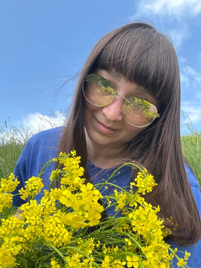 In the city of Sredneye Kechevo, Russia, a close-up shot captures a young woman with long, dark brown hair and bangs, wearing a blue t-shirt and reflective sunglasses. She is holding a large bouquet of small yellow flowers. The background consists of a bright blue sky with scattered white clouds and a field of tall green grass. The sunglasses reflect the yellow flowers and the sky.