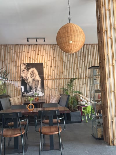 The image shows the interior of a cafe with a natural, rustic theme. The walls are adorned with vertical bamboo stalks, creating a textured backdrop. A large, woven spherical pendant light hangs from the ceiling, casting a warm glow. In the center of the room, there are several dark wooden tables with black-framed chairs featuring wooden seats. A striking black and white photograph of an elderly man smoking a pipe is displayed on the bamboo wall above one of the tables. Cushions with striped patterns are placed on seating areas. To the right, a glass display cabinet stands filled with decorative items. A potted plant adds a touch of greenery to the scene. The floor is a dark, tiled surface. The cafe is located in Abang, Indonesia.
