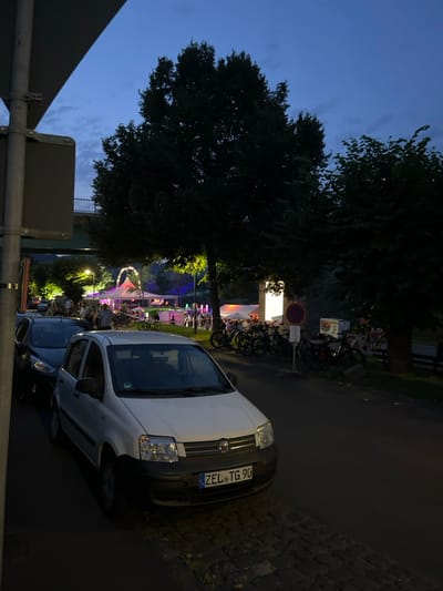This image captures an outdoor evening scene in Reil, Germany, characterized by the low light of dusk and illuminated event activity.
In the foreground, a white, four-door Fiat Panda hatchback, bearing German license plate ZEL TG 90 (identifying it as registered in the Cochem-Zell district, where Reil is located), is parked facing the viewer on a cobblestone surface. A dark-colored vehicle is partially visible parked behind it to the left. A dark utility pole with an obscured sign is positioned on the far left.
The midground features a line of parked bicycles and electric bicycles adjacent to a paved road. A red circular "no parking" sign with a diagonal line is visible near these bikes. Several large, dense trees partially obscure the background.
The background reveals a bustling outdoor event or festival, brightly illuminated with various artificial lights, including pink, purple, and blue hues. Multiple white tents or canopies are set up, with a prominent tall, white, arch-like structure or decorative piece visible towards the left side of the event area. Numerous individuals are seen moving and gathering among the tents and stalls, indicating active participation. A bridge or overpass is visible spanning overhead in the upper-left portion of the image, suggesting the presence of a road or natural feature beneath it. The sky is a dark twilight blue with faint cloud formations.