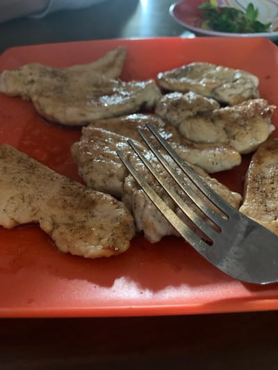 The image shows a collection of cooked chicken pieces on a red cutting board, with a metal fork resting on top. A plate with salad is partially visible in the upper-left corner. The location is not ascertainable from the image.