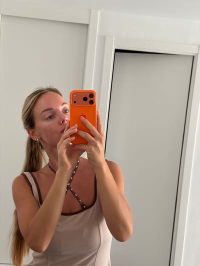 A young blonde woman is taking a selfie in a mirror. She is wearing a light-colored tank top with brown trim and crisscross straps with a polka dot pattern. She has her hair pulled back in a ponytail and is wearing earrings. She is holding an orange iPhone in her right hand. The background appears to be a well-lit room with white walls and trim. The overall setting suggests a personal space, possibly a dressing room or bathroom, within a residence. There is no visual information in the image that places the location specifically in Finestrat, Spain.