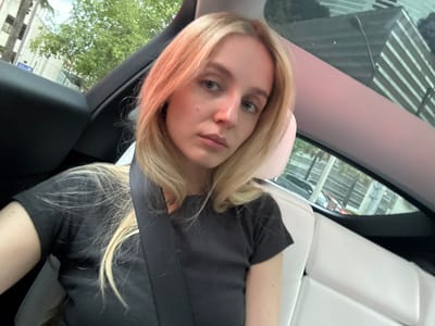 A young woman with blonde hair is seated in the back of a car. She is wearing a dark t-shirt and a seatbelt. The interior of the car appears to be white. Through the windows, a city environment is visible, with buildings and trees. The lighting suggests it is daytime.