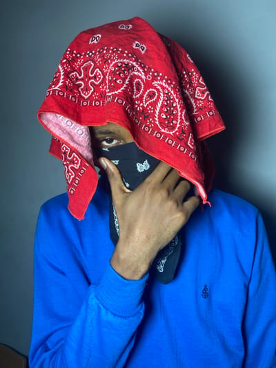 A person wearing a bright blue long-sleeved shirt is seen from the shoulders up. The person is facing left, with their face partially obscured by a red paisley bandana draped over their head and a black bandana covering the lower half of their face. Only one eye is visible. The person's right hand is raised to their face, holding the bandanas in place. The background is a solid gray.