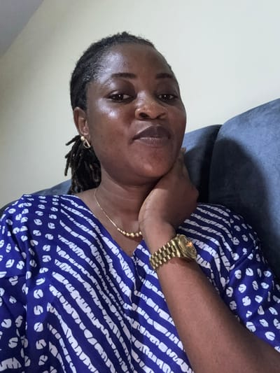 A medium shot shows a Black woman, likely in her 30s or 40s, with dreadlocks, looking directly at the camera. She is seated, with her left hand resting on her chin and her right arm visible, wearing a gold wristwatch. The woman has a gold necklace with round beads and gold earrings. She is wearing a blue and white patterned top. The background consists of a light-colored wall and a blue upholstered couch. The scene appears to be indoors, possibly in a residential setting in Ajara, Nigeria.