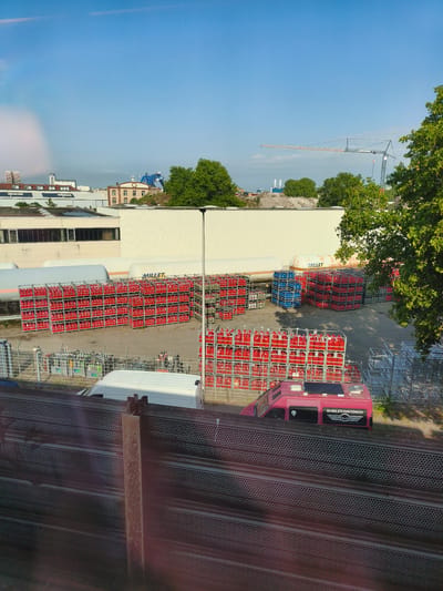 A high-angle, exterior shot captures an industrial area in Mannheim, Germany, under a clear blue sky. In the foreground, a metallic barrier with a perforated pattern runs horizontally across the frame. Behind this barrier, a white van is parked. Further back, a fleet of red gas cylinders, stacked in metal cages, dominates the scene. These are arranged in neat rows, with some empty metal racks to the right. A pink van, bearing a "HARLEY-DAVIDSON" logo on its side, is parked in front of some of the red cylinder racks. In the background, large white industrial buildings stand, with trees and construction cranes visible. The overall impression is one of industrial activity and storage.