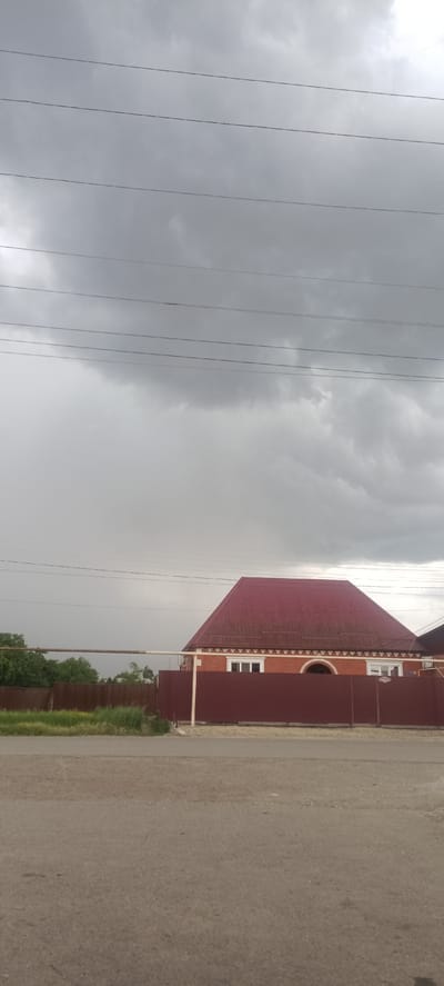 This is a landscape photograph taken from a low angle, looking towards a residential property in Ivanovskaya, Russia. The scene is dominated by a dramatic, overcast sky filled with heavy grey clouds, suggesting a storm is approaching or has recently passed. Five dark power lines are stretched horizontally across the upper portion of the frame, contrasting with the muted sky.

In the middle ground, a house with a prominent, steeply pitched red roof is visible. The house appears to be constructed of red brick and has several white-framed windows. A large, dark red metal fence encloses the property, with a section of green grass and some taller vegetation in front of it. A horizontal pipe, possibly part of a fence or utility structure, runs across the mid-section of the image. The foreground is a grey, asphalted road.

The overall impression is one of a quiet, possibly rural or suburban, setting under a brooding sky. There are no people visible in the frame. The time of day is difficult to ascertain precisely due to the overcast conditions, but the lighting suggests it is daytime, likely late afternoon or early evening. The weather is clearly unsettled. No visible text is present in the image.