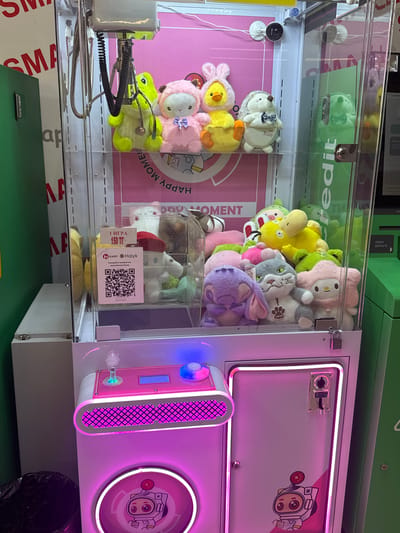 The image shows the interior of a crane game machine filled with plush toys, located in Kokshetau, Kazakhstan. The machine is pink and white with neon purple lighting around the bottom compartment. The crane mechanism is visible at the top. Plush toys include a pink sheep, a yellow duck, a hedgehog, a gray cat, a purple character resembling Stitch, and a pink bear. A QR code for payment is visible on a red and white sign, alongside text in Russian indicating "1 IGRA 150 TT" (1 game 150 Tenge). The background features a pink wall with "HAPPY MOMENT" printed on it. A green vending machine is partially visible to the right of the crane game.