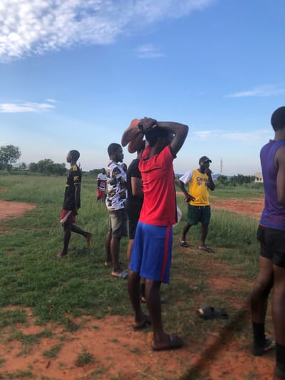 This image captures a group of young men and boys gathered in an open, grassy field under a clear blue sky with scattered white clouds. The setting appears to be late afternoon or early evening, characterized by bright, natural light. The ground consists of green grass interspersed with reddish-brown dirt paths.

In the foreground, a young man wearing a red short-sleeved shirt and blue shorts is holding a pair of brown sports shoes or cleats above his head, possibly engaging in a stretch or preparing for an activity. To his right, another man stands prominently in a yellow sleeveless shirt, which features the word "CHINA" in black letters and Chinese characters (likely "中国") beneath it. He is also wearing dark shorts and a baseball cap, and he looks towards the viewer with a casual hand gesture.

Behind these two, a man in a black and white patterned shirt and dark shorts is visible. Further left, a young boy in a black and red shirt with a partially visible logo (resembling Adidas stripes) and red shorts stands barefoot. Several other individuals are partially visible in the background and foreground, indicating a larger gathering. Sandals and other footwear are scattered on the ground near some participants. In the distance, low-rise buildings and trees suggest a semi-rural landscape. The overall atmosphere is casual, indicative of a sports practice, a game, or a social gathering in Nsugbe, Nigeria.