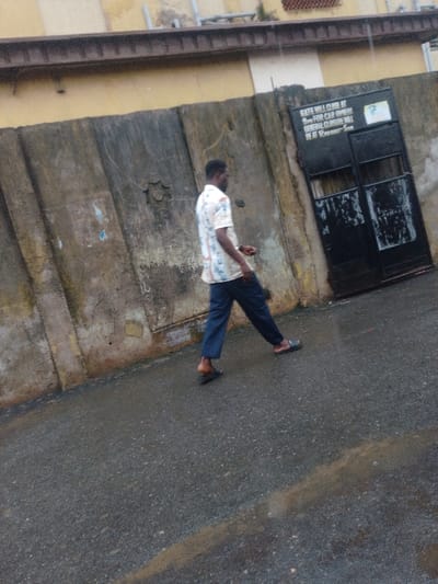 An adult male of dark complexion, wearing a light-colored patterned short-sleeved shirt, dark blue trousers, and dark open-toed sandals, walks from left to right across a wet, dark grey asphalt street in Agani, Nigeria. He is positioned in the mid-ground, with his right leg forward in motion. His right hand holds a small, dark object at waist level. To the left and behind the man is a weathered, light brown and grey concrete wall featuring vertical segments and a circular, star-shaped decorative element. Further right, a dark metal double gate is set into the wall. A white sign affixed to the upper left gate panel reads: "GATE WILL CLOSE AT 9PM FOR CAR OWNERS GENERAL CLOSURE WILL BE AT 12:00PM - 5AM". Above the wall, a section of a light yellow building with a dark horizontal structural element is visible. The street surface is visibly wet, indicating recent precipitation.