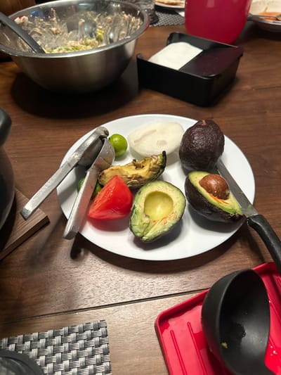 In Huixquilucan de Degollado, Mexico, a wooden dining table displays a spread of ingredients for a meal. A stainless steel mixing bowl, mostly empty, sits on the left, with traces of food residue inside. Nearby, a white plate holds chopped avocados, a tomato wedge, two small green limes, a slice of white onion, and a whole avocado. A metal lime squeezer rests on the plate among the ingredients. A knife with a black handle is also on the plate, next to a halved avocado. To the right of the plate, a black napkin holder contains white napkins. In the foreground, a red utensil rest holds a black ladle. The scene appears to be a preparation area for a meal, possibly guacamole given the avocados, tomato, and lime. The lighting suggests an indoor setting, likely a kitchen or dining room.