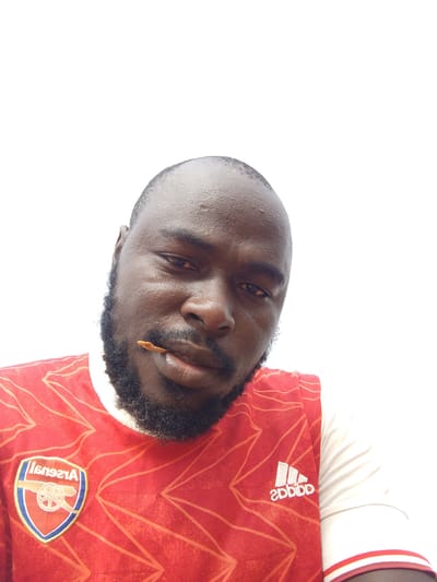 A selfie shows a Black man with a beard and closely cropped hair wearing a red and white Arsenal soccer jersey. He is holding a thin, brown object in his mouth. The Adidas logo is visible on his jersey. The Arsenal emblem is also visible. The background is white. The man appears to be looking down at the camera. Location information is not discernible from the image.