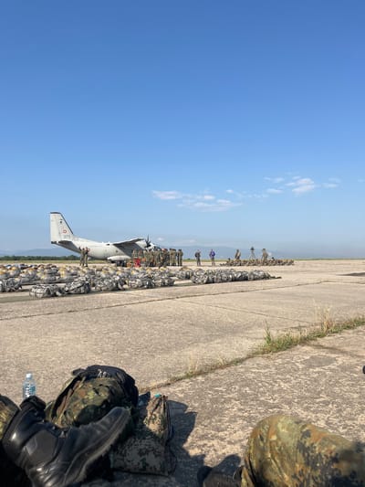 An outdoor, ground-level, wide shot captures an airfield scene under a clear blue sky with distant hills. In the foreground, military personnel sit on the tarmac, their legs visible, clad in camouflage uniforms and boots. Several large, camouflaged duffel bags and backpacks are arranged on the ground. A bottle of water is placed near a backpack. In the middle ground, a military transport plane, designated with the number "073" and bearing a circular emblem on its tail, is parked on the tarmac. A group of soldiers stands near the plane, some appearing to be loading or preparing equipment. Rows of duffel bags are neatly arranged on the tarmac in front of the aircraft. The background shows a treeline and rolling hills under the bright sun. The overall scene suggests preparation for a military operation involving air transport and parachuting. The location is the airfield in Cheshnegirovo, Bulgaria.