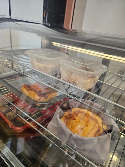 A refrigerated display case at a food establishment in Banyuurip, Indonesia, contains several food items. On the top shelf, two clear plastic containers hold food that appears to be either pasta or rice dishes. The containers are slightly fogged with condensation. On the shelf below, a rectangular glass dish holds a piece of cooked meat, possibly pork belly, with a golden-brown, slightly charred crust. Next to it, on the bottom shelf, is a round baked item, likely a dessert such as a cheesecake or tart, lined with parchment paper and presented in a metal tin. The display case is lit from above, creating reflections on the glass exterior. The environment appears clean and orderly, typical of a commercial food service setting.