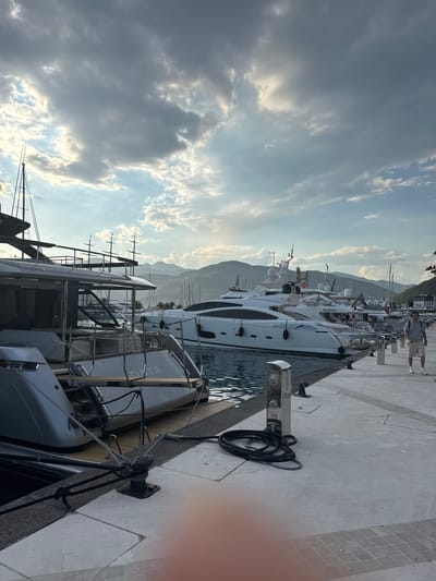 A man in shorts and a t-shirt walks along a paved pier in Tivat, Montenegro, past several docked yachts. The yachts are white and sleek, with dark trim and masts. The sky is cloudy with patches of sunlight. In the background, mountains rise behind the town. The scene appears to be in the Porto Montenegro marina, a luxury yachting hub in Tivat.