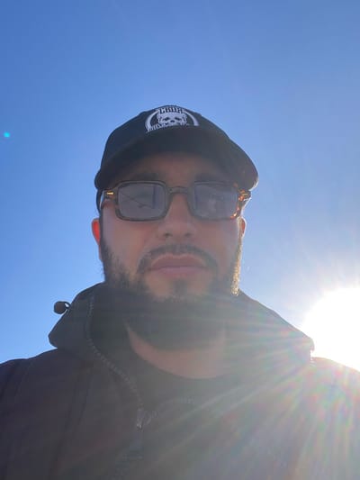 The image is a selfie taken outdoors against a bright blue sky, with the sun shining intensely and causing lens flare. The subject is a man wearing a black baseball cap with a white skull logo and patterned sunglasses. He has a beard and is wearing a dark jacket. The setting appears to be a sunny day, possibly with clear weather. There is no indication of the location being Koskolovo, Russia, or any specific urban environment.