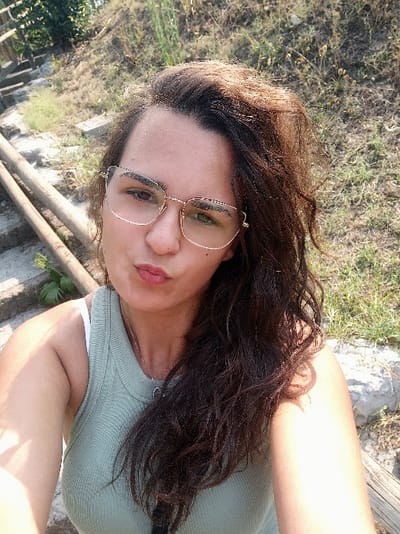 The image captures a single adult female in an outdoor setting in San Marzano sul Sarno, Italy. The subject is positioned in the foreground, facing the camera directly, indicating a self-portrait. She possesses dark, wavy hair and wears gold-framed eyeglasses and a light green ribbed tank top over a white strap. A silver-toned necklace with a circular pendant hangs from her neck. Her lips are pursed, and her gaze is directed towards the lens. The background on the left reveals a partial view of a stone staircase ascending uphill, flanked by two parallel wooden handrails. The background on the right consists of dry, sparse grass and scattered vegetation on an upward incline. The scene is illuminated by bright, natural daylight.