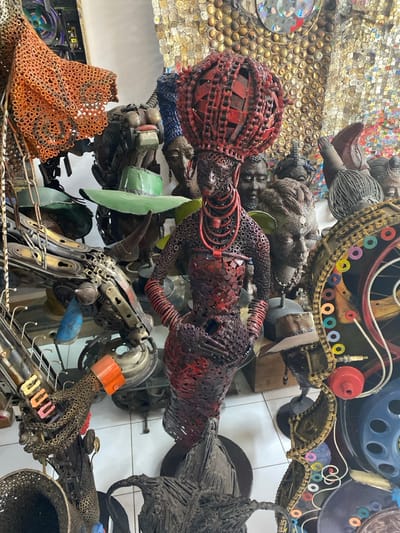 The image displays an interior view of an art space or workshop in Alaguntan, Nigeria, densely filled with sculptures constructed from reclaimed metal components. The central subject is a life-sized, standing female figure, meticulously assembled from nuts, bolts, chains, and sheet metal, painted predominantly in red and black. She wears a prominent, spherical red headdress, multi-strand red necklaces, and red armbands, with her hands positioned on her midsection. The surrounding area is populated by numerous other metallic sculptures, including busts and abstract forms crafted from similar industrial waste. Notable background elements include a bust adorned with a green wide-brimmed hat, intricate assemblages of bicycle parts and other metallic objects on the left, and a large wall-mounted mosaic composed of metallic bottle caps and reflective discs. The floor of the space is white-tiled.
