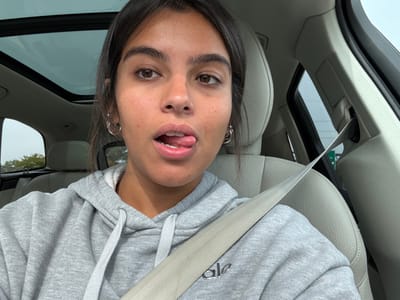 A female individual is depicted in the passenger seat of a vehicle in West Windsor, United States. She has dark hair and brown eyes, and wears small hoop earrings. Her mouth is open with her tongue partially extended. She is dressed in a grey hooded sweatshirt with the text "ale" visible on the front, and is secured by a light-colored seatbelt. The vehicle interior features light-colored seating and a panoramic sunroof reflecting an overcast sky. Green foliage is visible through the left window, and a green road sign or structure can be seen through the right rear window.