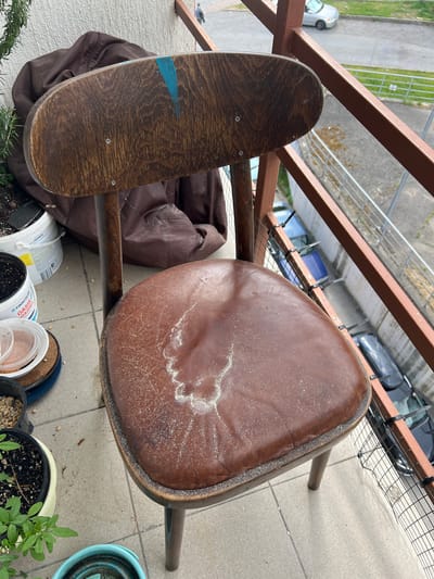 An outdoor balcony in Krasnaya Polyana, Russia, features a weathered wooden chair with a brown leather seat that has a powdery white residue scattered across it. The chair has a curved wooden backrest with a vertical blue stripe painted near the top. Behind the chair, a dark brown fabric object, possibly a jacket or bag, is visible. Various potted plants and containers are placed on the tiled balcony floor. In the background, a metal railing overlooks a street with parked cars and a building.