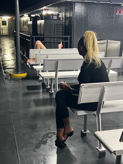 The image captures a nighttime scene on what appears to be the deck of a ferry or transport boat, likely in the city of Nathon, Thailand, given the context. In the foreground, a woman with blonde hair in a high ponytail is seated, facing away from the camera and to the right. She is wearing a black outfit and black sandals, with a turquoise anklet visible on her right ankle. She holds a white paper cup with a straw in her left hand and appears to be adjusting her shoe with her right hand. Adjacent to her, a man's legs and feet are visible, stretched out on a white bench or seat, suggesting he is lying down or reclining. He is wearing khaki shorts. The deck is dark and appears wet, with reflections from overhead lighting. Rows of white, metal-framed benches line the deck. In the background, a white door with a railing is visible, along with darkened windows and structures suggesting the interior of the boat. The overall atmosphere is dimly lit, with artificial lights casting shadows.