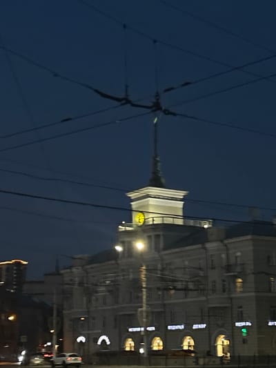 The image depicts a street scene at dusk in Barnaul, Russia. Dominating the upper portion of the frame are numerous overhead wires, likely for public transportation, crisscrossing the dark blue sky. Below the wires, a distinctive building with a tower featuring a clock is visible. This structure appears to be a prominent landmark. The building's facade is adorned with multiple windows, some illuminated, suggesting indoor activity.
In the lower part of the image, several vehicles are present on the street, with headlights and taillights indicating movement or stationary positions. The street itself appears to be paved. The overall environment suggests an urban setting during the evening hours, with artificial lights illuminating parts of the street and buildings. The buildings in the background, though less distinct, reinforce the urban context. The presence of streetlights and illuminated windows contributes to a sense of a city that is active even as daylight fades. The image captures a moment in time on a street in Barnaul, Russia, characterized by its infrastructure and architecture under the twilight sky.