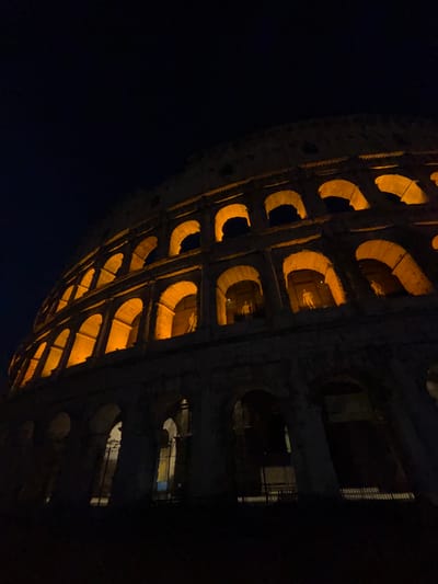 The image captures a low-angle, wide shot of the Colosseum in Rome, Italy, at night. The ancient amphitheater is illuminated by warm, orange lights, highlighting its iconic arched structure. The sky above is a deep, dark blue, indicating nighttime. Several arches on different levels are visible, with some of them containing dimly lit figures that appear to be statues or mannequins. The lower sections of the Colosseum are in deep shadow, with only faint outlines of arches and openings visible. In the foreground, at the very bottom of the image, there are dark, shadowed areas that suggest the ground level or lower structural elements. The overall impression is one of grandeur and historical significance, with the lighting emphasizing the architectural details of the monument against the night sky.