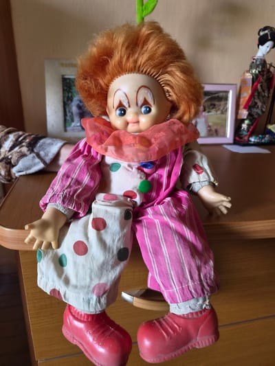 A vintage doll, dressed as a clown, is seated on a wooden surface. The doll has bright orange, curly hair and a round, plastic face with blue eyes and rosy cheeks. It wears a pink and white polka dot outfit with striped pink pants and a ruffled collar. Red clown shoes complete the costume. Behind the doll, on a shelf, are two framed pictures and a small Japanese doll in a kimono. The background appears to be a wall, suggesting an indoor setting. This scene is located in the city of Arseniev, Russia.