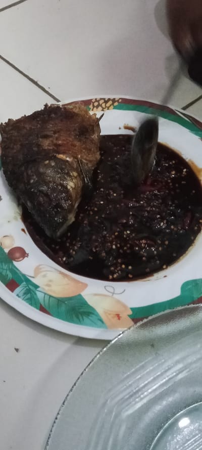 A white ceramic plate with a green and brown floral design sits on a white tiled counter. On the plate is a piece of fried fish and a dark brown sauce. The fish is dark brown and has a crispy texture. The sauce has small white and red specks in it. A large metal spoon is partially submerged in the sauce. Another plate, made of clear glass with a raised lip, is partially visible to the lower right of the ceramic plate. The scene takes place in Cangkuang, Indonesia, at a location with coordinates latitude -7.065 and longitude 107.552.