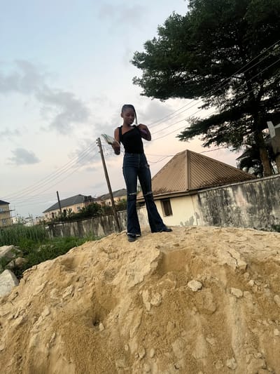 A young individual, appearing female, stands atop a large mound of sand in Iranla, Nigeria. She wears a black sleeveless top, dark blue flared jeans with distressed areas, and dark shoes. Her hair is styled in braids. In her left hand, she holds a small book or notebook with a visible green cover, while her right hand is raised towards her head. The sky above is overcast with light grey and white clouds.
In the mid-ground, multiple utility poles with strung electrical wires traverse horizontally. To the right, a large tree with dense green foliage extends into the upper frame. Behind the sand mound, a concrete perimeter wall, weathered and discolored, is visible, topped with a coil of barbed wire on the right side. Behind this wall, a light-colored, single-story building with a brown corrugated metal roof is present. Further to the left in the background, other residential-style buildings with dark roofs and light-colored walls are visible, one appearing to be a multi-story structure. Green vegetation is interspersed between the sand pile and the wall. The foreground consists entirely of the granular, light-brown sand mound, showing uneven surfaces and small depressions.