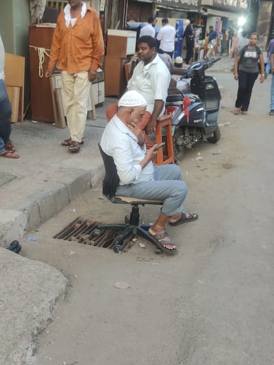 A street scene in Mumbai, India shows an elderly man sitting on an office chair positioned directly over a sewer grate. He is fair-skinned and wearing a white topi, a white collared shirt, grey trousers, and sandals, and he is looking at a cell phone in his hand. Behind him is a second, darker-skinned man wearing a white collared shirt and sitting on a small wooden stool next to a black scooter with the words "Easygo One" written on it. In the background are pedestrians, storefronts, and various goods, suggesting a commercial area. The street is made of concrete and shows signs of wear.
