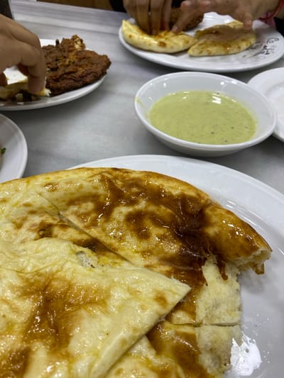 The image captures an indoor dining scene in Narayanganj, Bangladesh. In the foreground, a white oval plate displays multiple pieces of glossy, golden-brown naan bread, with one piece partially torn. Centered in the midground is a small white bowl containing a light green, textured sauce. Further back on a light grey table, two additional white plates are visible. The plate to the left holds a large, dark brown, fried meat item, from which a hand is tearing a portion. The plate to the right contains two pieces of naan, and another hand is reaching towards them. The background is out of focus, indicating an active restaurant environment.