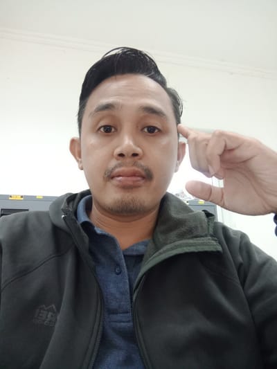 A medium-shot photograph shows a man wearing a dark-colored jacket and a blue collared shirt. His hair is dark and styled upwards. He has a mustache and light stubble. His right hand is raised, with his index finger near his temple, as if he is thinking. The background is a plain white wall, with a hint of cabinets behind the left shoulder. The man faces the camera with a neutral expression.