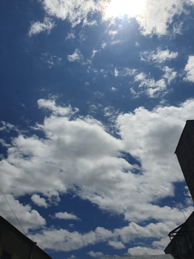 A low-angle shot captures a bright blue sky filled with fluffy white clouds. The sun is visible on the left side of the frame, casting rays of light that create a lens flare effect. The clouds are scattered across the sky, with some appearing dense and cumulus-like, while others are wispy and streaky. The bottom right and top portions of the image show the edges of buildings, suggesting an urban or semi-urban setting. The buildings have dark gray, textured facades and the rooflines are visible. A thin black wire runs diagonally across the lower right corner, attached to one of the buildings. The overall scene suggests a clear day with a mix of sun and clouds. Given the location context of Gavar, Armenia, this could be a typical daytime sky in the region. There is no visible text in the image.