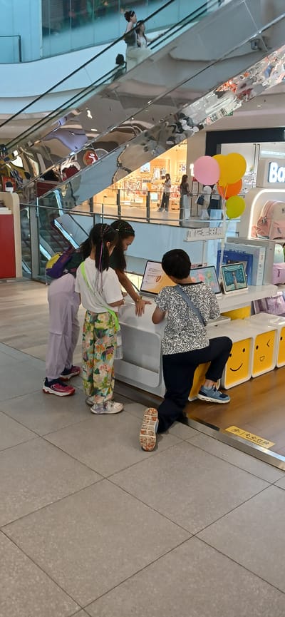 In a brightly lit indoor retail space, possibly a department store or a dedicated brand store in Jiulongpo District, China, four individuals are gathered around a display counter. The scene is characterized by modern retail design elements, including a sleek, reflective escalator in the background and a white counter unit adorned with display screens and colorful cubic storage boxes featuring smiley face designs.

Three young individuals, appearing to be children, are actively engaged with the display. Two girls, standing on the left, are looking at a tablet or interactive screen on the counter. The girl in the foreground has her hair styled with colorful braids, and she is wearing a white t-shirt and floral patterned pants. To her left, another girl in a dark top and light purple pants, with a backpack on, also seems to be observing the screen. To the right, a third child, dressed in a patterned t-shirt and dark pants, is kneeling down, also interacting with the display.

Behind the counter, several other individuals are visible, including two women ascending an escalator, their reflections caught in the polished surface. The overall environment suggests a commercial setting designed to attract and entertain shoppers, with the presence of balloons adding a festive touch. The floor is tiled in a neutral color. The retail space features product displays, including backpacks, and branding that is partially visible.