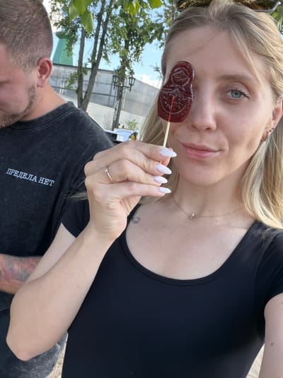 A young blonde woman with fair skin is holding a red, matrioshka-shaped lollipop in front of her right eye, looking at the camera. She is wearing a black t-shirt and a small, delicate necklace. Her nails are painted white. To her left, a man with short brown hair and a goatee is partially visible, wearing a dark t-shirt with white Cyrillic text that reads "ПРЕДЕЛА НЕТ". They appear to be outdoors, with trees and a partially obscured building in the background. The overall atmosphere suggests a casual outing in Suzdal, Russia.
