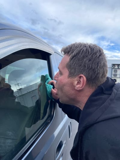 A man is cleaning the window of a dark-colored vehicle. He is holding a blue cloth and appears to be wiping the glass. The reflection in the window shows a building. The sky is cloudy. The location is Бавыкино, Russia.