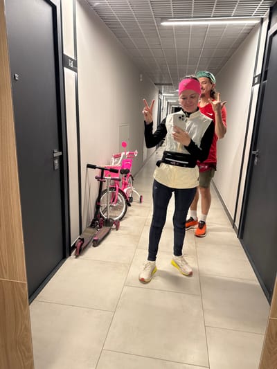 Two individuals stand in an indoor hallway. The first individual wears a pink headband, black long-sleeved shirt, beige jacket, black leggings, and white running shoes with yellow soles. They hold a phone and make a peace sign. The second individual, behind them, wears a green and black baseball cap, red shirt, and green shorts, also making a hand gesture. A pink bicycle and scooter rest against the wall beside the first individual. The hallway has grey doors, tiled floors, and fluorescent lighting. The image was likely taken in Verkhnii Liuk, Russia.