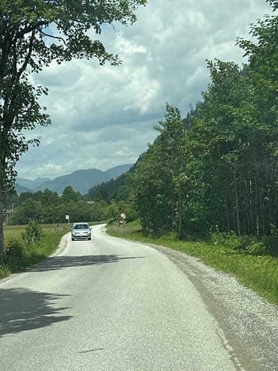 The image shows a view from a vehicle traveling on a paved road through a rural, mountainous landscape. The road curves to the right, with a white car visible in the distance ahead, driving in the same direction. The left side of the road is bordered by a dense line of trees, with a forest extending up the side of a mountain. Beyond the trees, layers of mountains are visible, fading into the distance under a cloudy sky. The right side of the road has a gravel shoulder and grassy verge, with more trees and vegetation. Sunlight casts long shadows from unseen trees onto the road. A circular sign with red markings and what appears to be a number is visible on the right side of the road. Another small white sign is posted near the car. The scene suggests a daytime drive through a scenic, possibly alpine, region in Austria, under overcast or partly cloudy weather conditions.