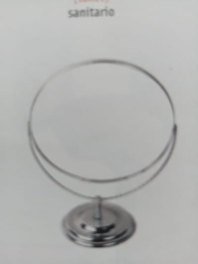 A close-up, slightly blurred photograph shows a round, tabletop mirror with a silver metal frame and base, set against a plain white background. The word "sanitario" is visible in small, grey text at the top of the image, suggesting the mirror is for sanitary use. The mirror is angled slightly upwards. The lighting is soft and even, with no harsh shadows. There are no people or other objects visible in the frame. The photograph does not contain any information about the location in the city of La Tagua, Venezuela.