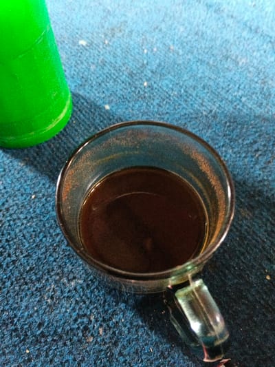 An overhead view depicts a clear glass mug containing dark brown liquid, likely coffee, positioned on a textured, light blue surface in Bien Hoa, Vietnam. The inside rim of the mug exhibits a light brown residue. A bright green, cylindrical object, possibly a plastic bottle or cup, is partially visible and out of focus in the upper left quadrant of the frame. The blue surface shows scattered white particulate matter. No individuals, actions, or interactions are visible.